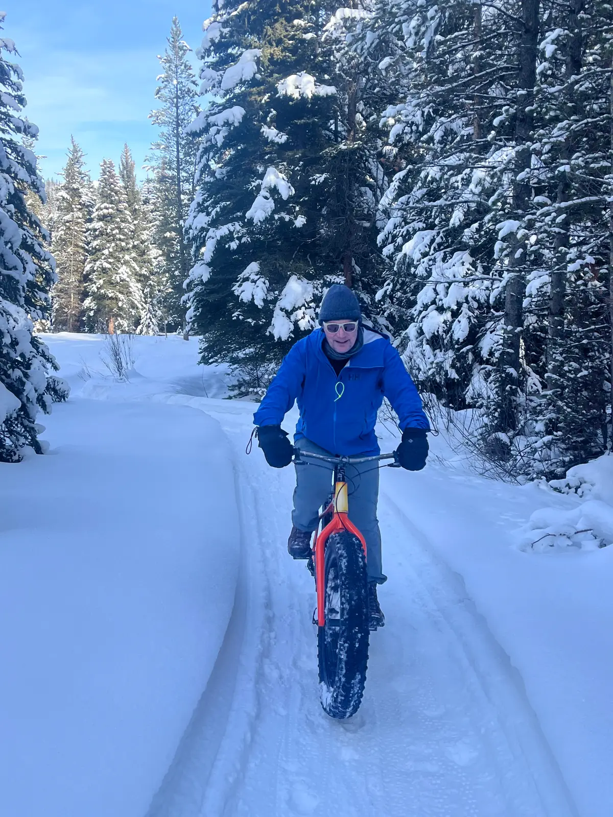 A rider on a fat-tire bike coming up a packed snow trail through heavily snow-laden evergreens