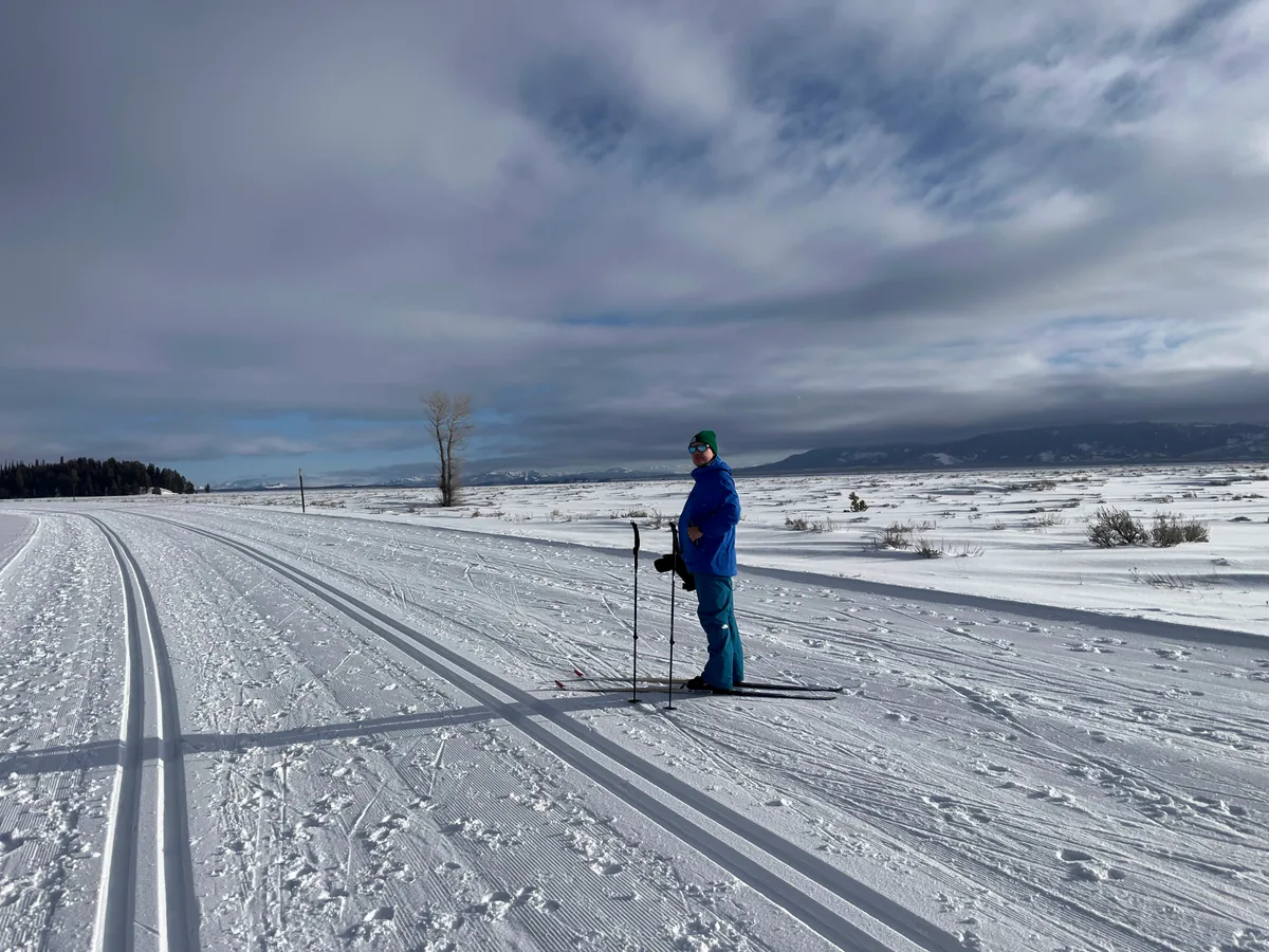 A cross-country skier in a blue jacket pausing on groomed classic tracks across an open snowy plain under heavy cloud