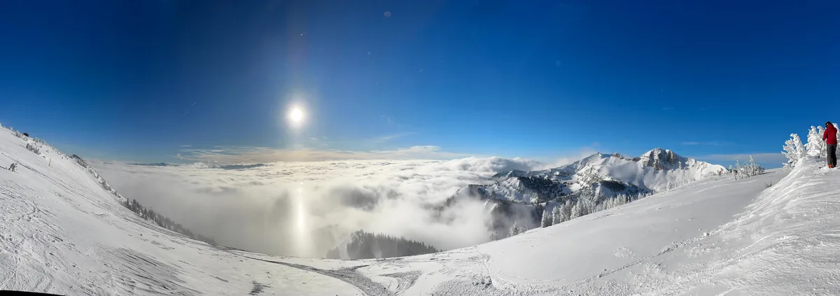 Panoramic view from a groomed ski run above a valley filled with cloud, snow-covered peaks catching the sun beyond