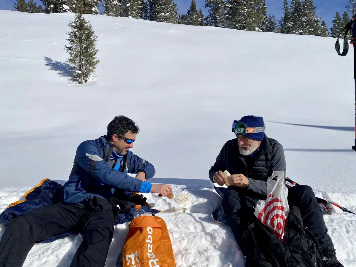 Two skiers sharing lunch while seated in deep snow beside a mountain trail