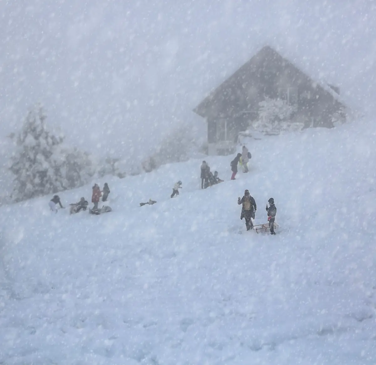 Heavy snowfall over a timber lodge with people sledding on the slope below