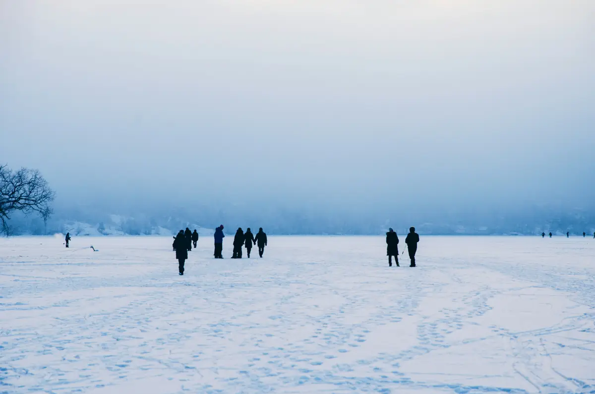 Distant figures crossing a vast flat snowfield as fog closes over the far treeline