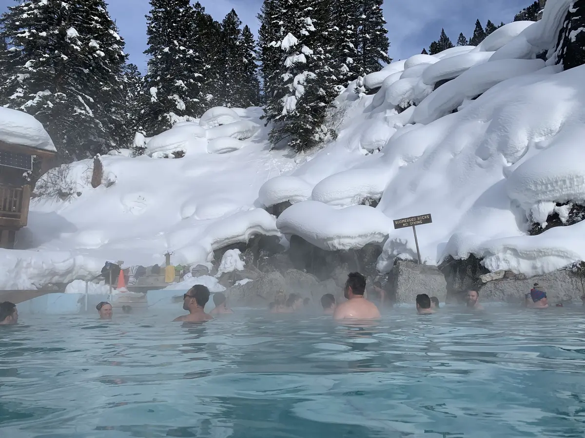 Travelers soaking in an outdoor hot-springs pool surrounded by deep snow and pine trees