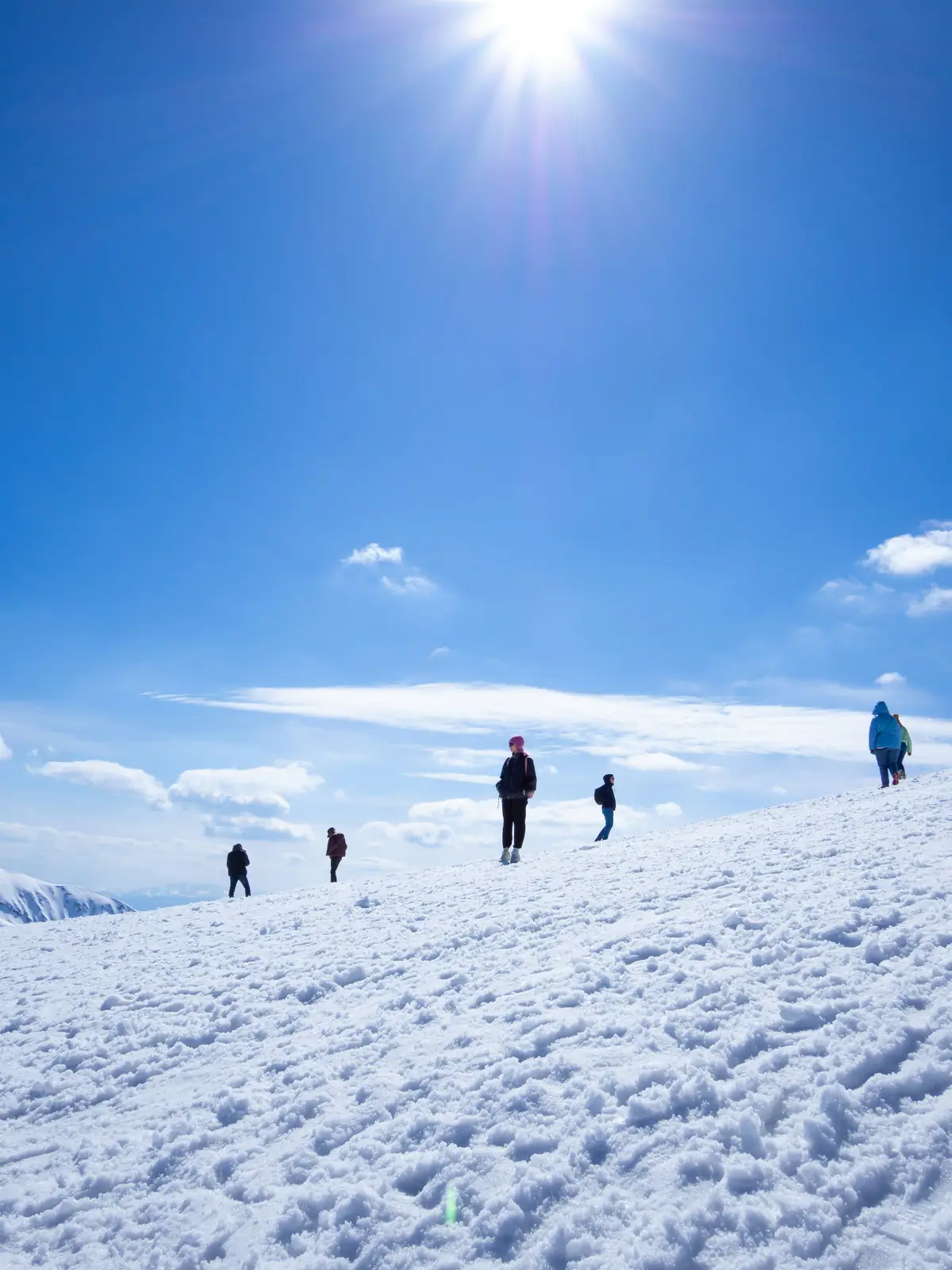 Small figures spread across a bright snow slope climbing toward the sun above a bank of cloud