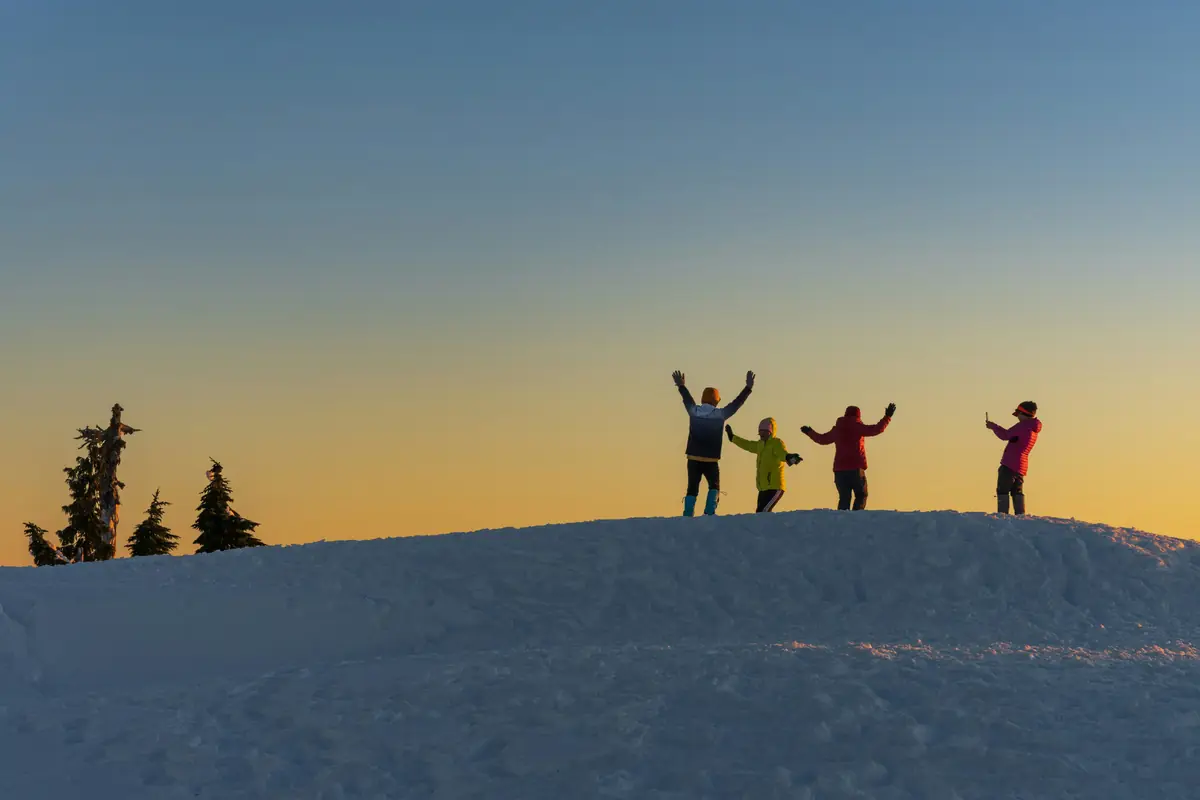 Four figures on a snowy ridge with arms raised against an orange and blue sunset sky