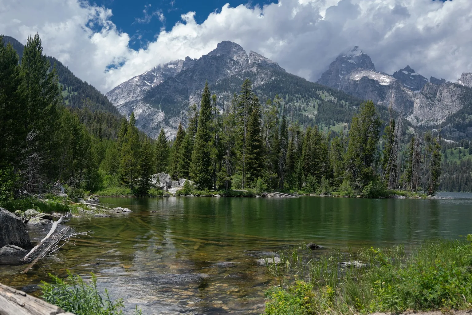 A clear alpine lake ringed by pines below the Teton range