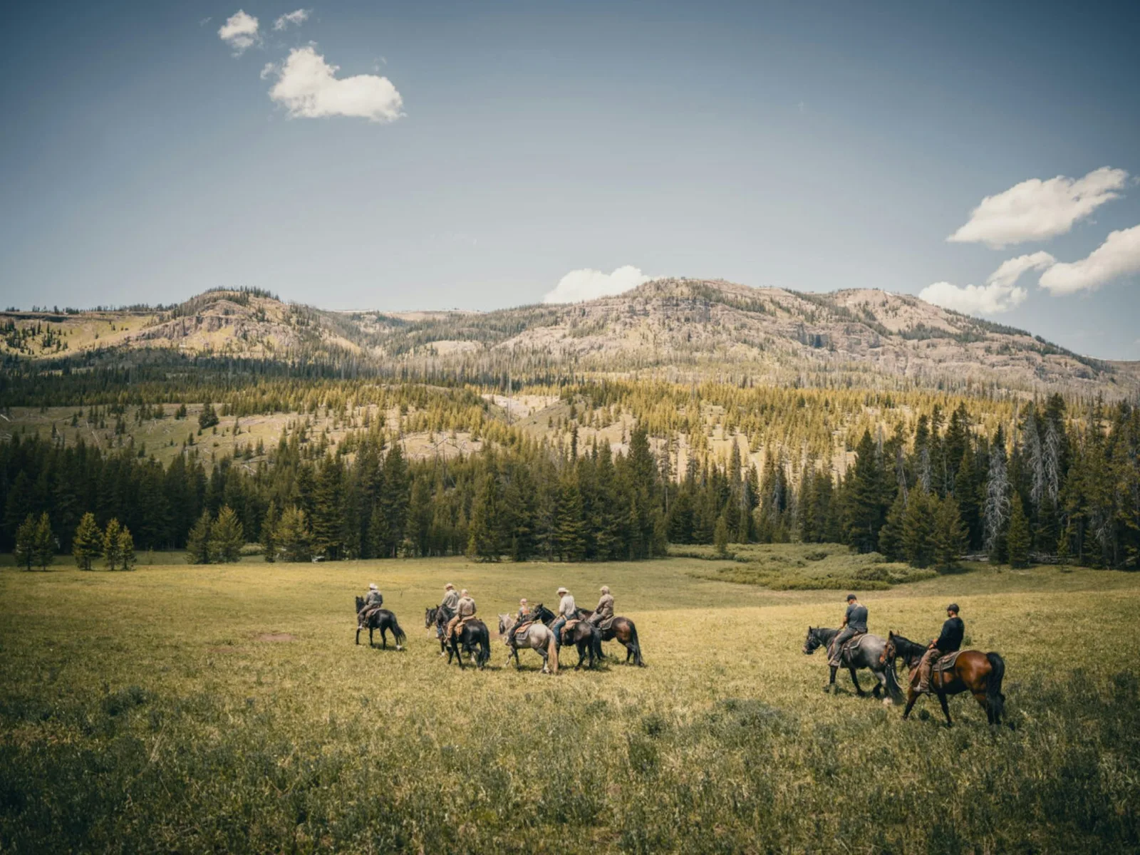 A group of riders on horseback crossing an open meadow below forested ridges